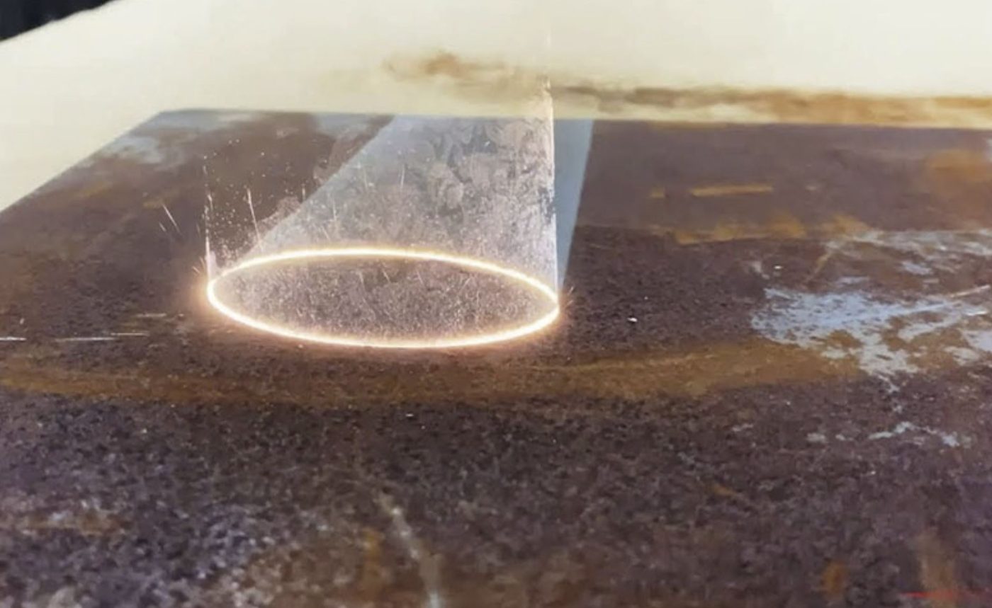 Laser beam stripping rust from a steel plate, leaving a clean bare-metal ring
